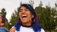 a woman with blue and purple hair and hoop earrings is laughing with her mouth open .