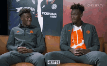 two men sit on a couch in front of a yokohama tyres sign