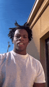 a man with dreadlocks is wearing a white t-shirt and smiling while standing in front of a building .