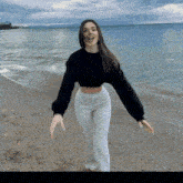 a woman wearing a black sweater and white pants is standing on a beach