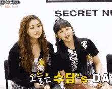 two girls sit next to each other in front of a sign that says secret