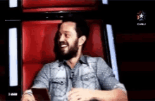 a man with a beard is sitting in a red chair laughing while holding a cell phone .