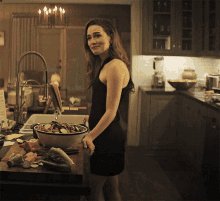 a woman in a black dress is standing in a kitchen with a bowl of food on the counter