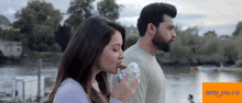 a man and a woman are standing next to a body of water and the woman is eating a donut