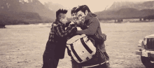 three men are hugging each other in a field while a jeep is parked in the background .