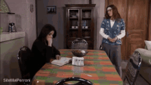 a woman sits at a table reading a book while another woman stands behind her and talks to her