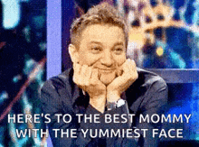 a man is smiling with his hands on his face and says here 's to the best mommy with the yummiest face .