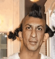 a man with a mohawk and pigtails on his head is standing in front of a window .