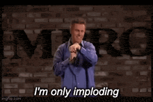 a man in a blue shirt is holding a microphone and saying i 'm only imploding