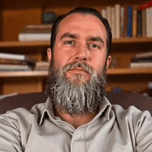 a man with a beard is sitting in a chair in front of a bookshelf and looking at the camera .