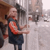 a bald man in a blue and orange jacket is standing on a sidewalk holding a pole