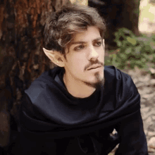 a man wearing elf ears and a black cape is sitting next to a tree