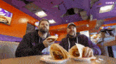 two men are sitting at a table eating tacos in a restaurant with a luma logo on the ceiling
