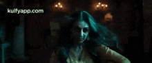 a woman in a yellow shirt is standing in a dark room with the website kulfyapp.com in the corner .