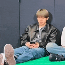 a man in a leather jacket is sitting on the floor looking at his cell phone