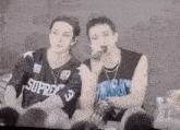 a couple of men are sitting next to each other on a stage . one of the men is holding a microphone .