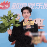 a man in a black suit holds a plant in front of a sign that says sina