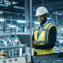 a man wearing a hard hat and safety vest is using a tablet computer in a factory .