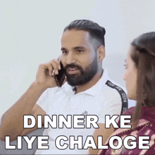 a man with a beard is talking on a cell phone while a woman watches .