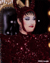 a drag queen is sitting in a chair wearing a red sequined dress and a devil costume .