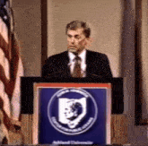 a man in a suit and tie stands at a podium with a shield on it that says cumberland university