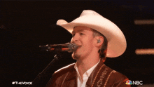 a man in a cowboy hat singing into a microphone with the nbc logo behind him