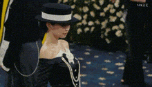 a woman wearing a hat and a necklace of pearls is standing on a blue carpet .