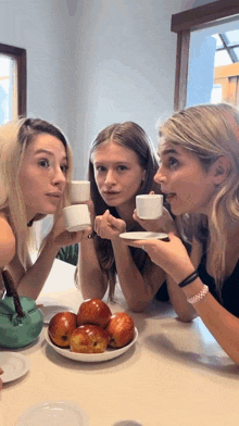 three women are sitting at a table holding cups of coffee