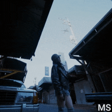 a woman in a leather jacket stands in a narrow alleyway looking up at a plane in the sky