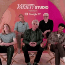 a group of men are sitting in director 's chairs in front of a variety studio sign .