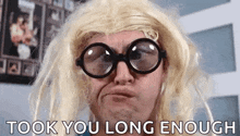 a man wearing glasses and a blonde wig is making a funny face and saying `` took you long enough '' .