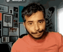 a man with a beard wearing a red shirt is sitting in front of a wall with pictures on it .