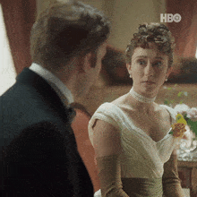 a man in a tuxedo and a woman in a white dress are looking at each other with the hbo logo in the corner