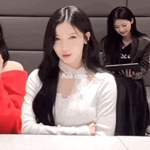 a woman is sitting at a table with her arms crossed and looking at the camera while another woman looks on .