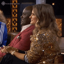 a man and a woman are sitting on a couch . the woman is wearing a gold sequined jacket .