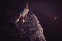 a woman in a white tank top and white shorts stands on a beach at night
