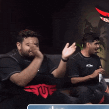 two men are sitting on a couch and one of them is wearing a shirt with the letter u on it