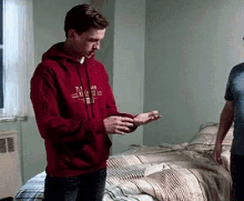 a man in a red hoodie is standing in front of a bed and looking at his phone