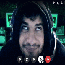 a man wearing headphones and a hoodie looks at the camera with a green screen behind him that says bu