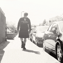 a man wearing a kilt is walking down a street with cars parked behind him