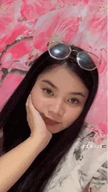 a woman wearing sunglasses looks at the camera in front of a pink flower background