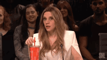 a woman is drinking from a red cup with a straw while sitting in front of a snl logo