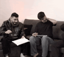 two men are sitting on a couch one is holding a tablet and the other is holding a piece of paper