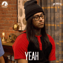 a man with long hair and glasses is wearing a black beanie and a red shirt that says yeah