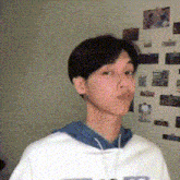 a young man wearing a white shirt and a blue hoodie stands in front of a wall with pictures on it