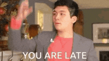a man in a suit and red shirt is saying `` you are late '' while waving his hand .