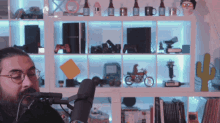 a man wearing glasses stands in front of a microphone in front of a shelf full of toys