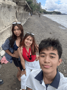 three people posing for a picture on a beach one of the girls is wearing a red top