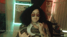 a woman wearing sunglasses stands in front of a wall that says happy halloween