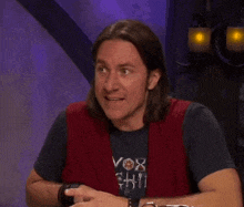 a man with long hair is sitting at a table with his mouth open and wearing a vox chili shirt .